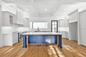 Modern kitchen with white cabinets, a large central island with a blue base, stainless steel appliances, and hardwood floors. Designed by a Dallas custom home builder, natural light enters through a wide window above the sink.
