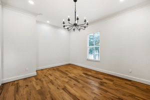 Empty room with white walls, hardwood floor, a window with blinds, and a black chandelier—showcasing the elegance you can expect from a Dallas custom home builder.