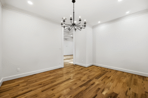 Empty room with white walls, a black chandelier, recessed ceiling lights, and hardwood flooring, showcasing the elegance possible with custom home builder in Dallas craftsmanship and an open doorway leading to another room.