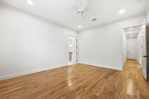 Empty room with light-colored walls, wooden floor, white ceiling fan, recessed lighting, and open doorways leading to a bathroom and hallway—crafted by a Dallas custom home builder for your dream build.