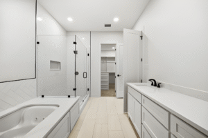 Modern bathroom by a Dallas custom home builder, featuring a glass-enclosed shower, built-in bathtub, double sink vanity, and a walk-in closet with white shelves in the background.