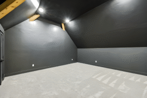 A dark gray, empty attic room with sloped ceilings, exposed wooden beams, recessed lighting, and clean, light-colored carpet—crafted with the quality you’d expect from a Dallas custom home builder.