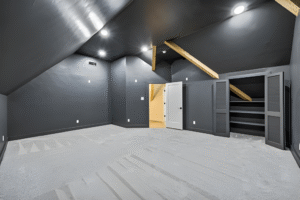 A finished attic room with dark walls and ceiling, light carpet, exposed beams, recessed lighting, open closet, and an open door leading to a lit hallway—expertly crafted by a Dallas custom home builder.