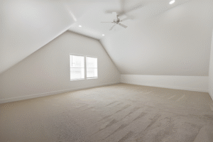 Spacious empty attic room with beige carpet, white walls, two windows, sloped ceiling, ceiling fan, and recessed lighting—perfect for your custom design by a leading Dallas custom home builder.