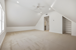 Spacious, empty attic room with beige carpet, white walls, built-in shelves, a ceiling fan, recessed lighting, and two doorways. Natural light enters from a window on the left—crafted by a Dallas custom home builder for comfort and style.