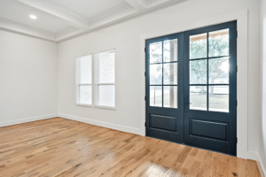 Bright, empty room with light wood floors, white walls, two shuttered windows, and double black French doors with glass panes letting in natural light—an ideal space crafted by a custom home builder in Dallas.