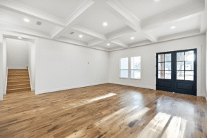 Bright, empty living room with coffered ceiling, hardwood floors, large windows, double front doors, and a staircase—crafted by a leading Dallas custom home builder for refined elegance and timeless style.