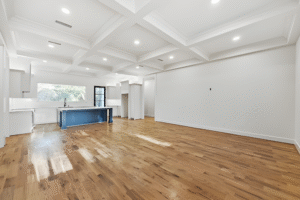Spacious open-plan room with hardwood floors, white walls, coffered ceiling, recessed lighting, and a modern kitchen featuring a blue island and large window—crafted by a top Dallas custom home builder.