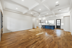 Spacious, empty open-concept room with wood floors, white walls, a coffered ceiling, and a kitchen area featuring a large island—crafted by a custom home builder in Dallas for modern living.