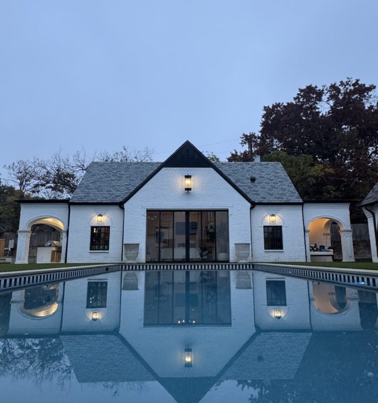 A white house with a gray roof, remodeled by About Custom Coats Remodeling, is reflected in a still swimming pool on a cloudy day, with trees and overcast sky in the background.
