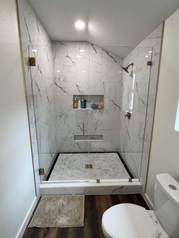 Modern bathroom with a large glass-enclosed shower featuring marble walls, gold fixtures, a built-in shelf, and a white toilet beside a beige bath mat on wood flooring—perfect inspiration for Bathroom Remodeling Dallas.